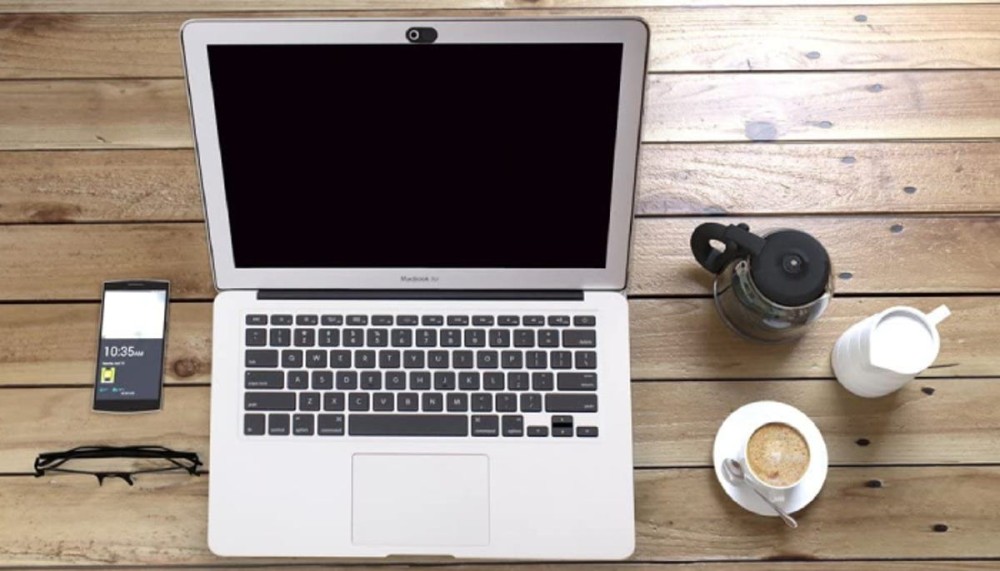 silver laptop with a black camera cover, sitting a wooden plank table next to glasses, a smartphone, and coffee cups