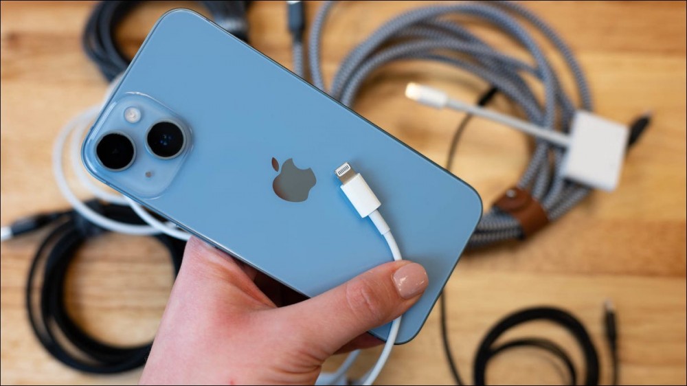 Person's hand holding an iPhone with a Lightning cable, and more cables in the background.