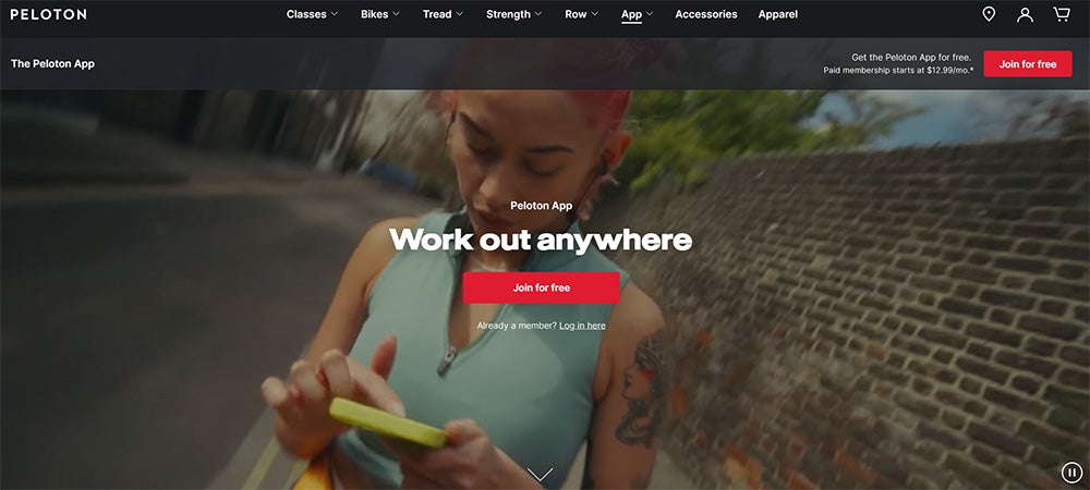 A woman dressed in workout clothes outdoors swiping on her smartphone on the Peloton app website.  