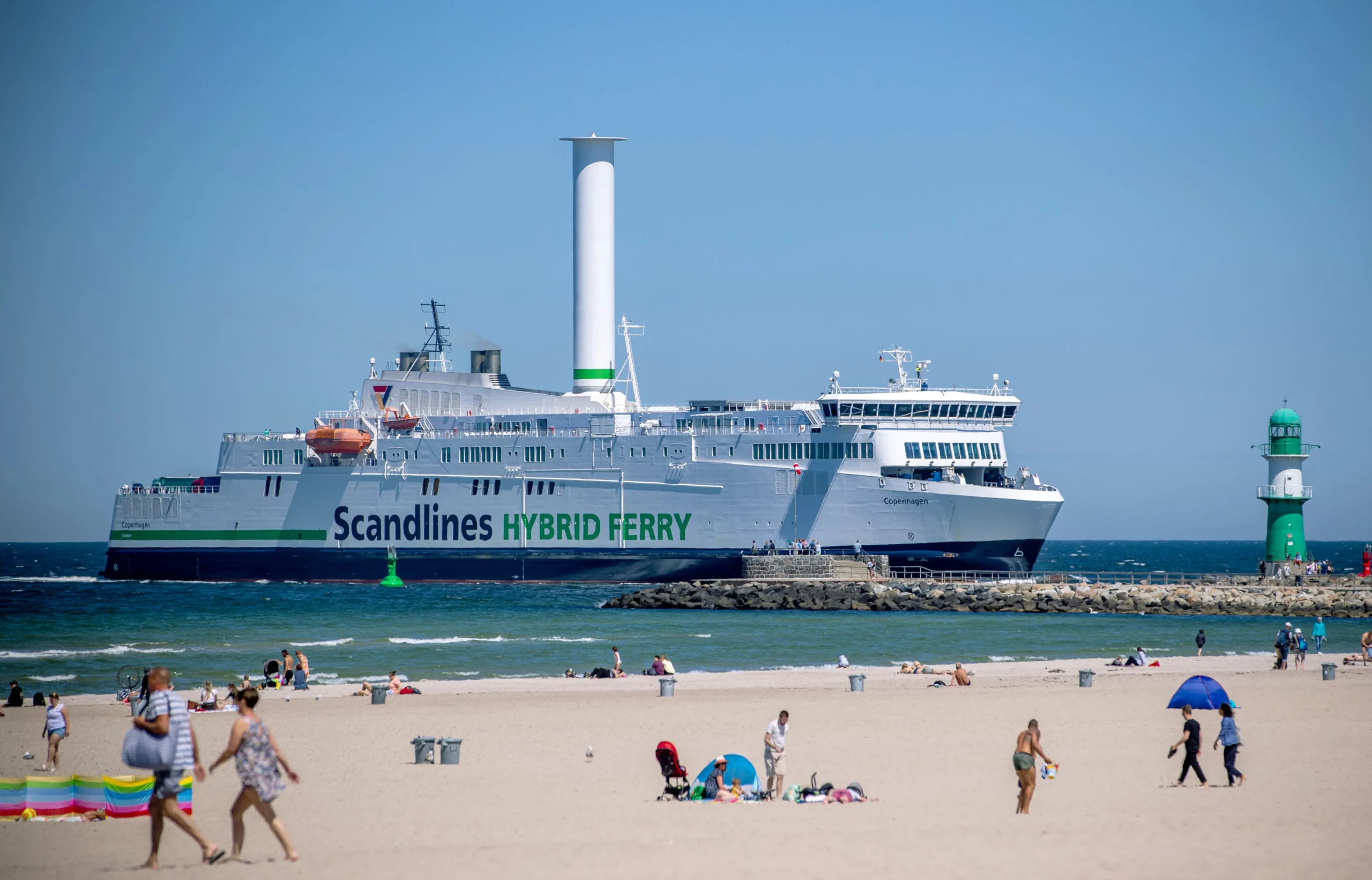 A color photograph shows a large white hybrid ferry with "Scandlines" and "HYBRID FERRY" written on its side. The ferry has a tall, white, spinning rotor sail on its deck. It is moving across a blue sea near a sandy beach with people sunbathing and walking, and a green lighthouse is visible in the distance. The sky is clear and blue.