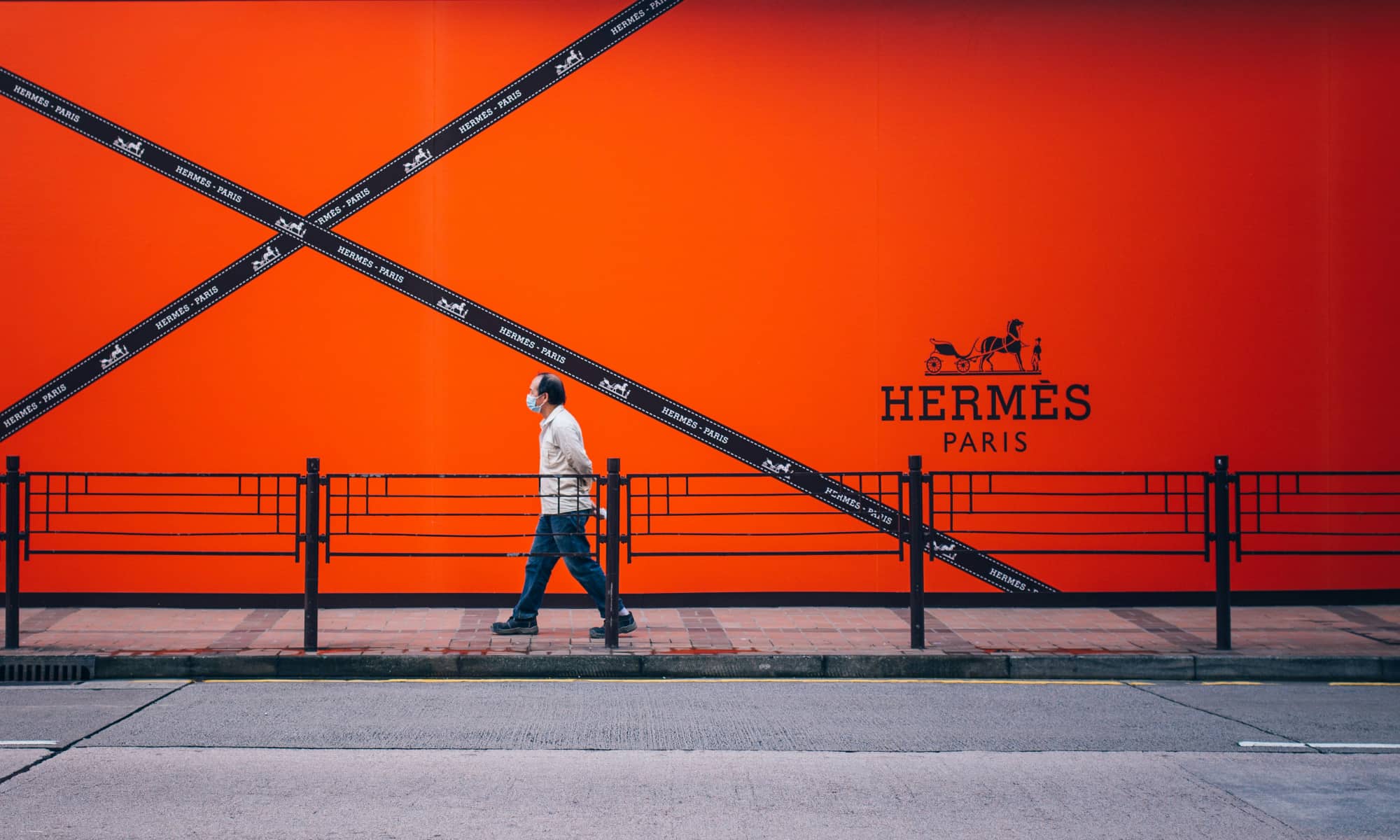 A man wearing a face mask walks past a massive, vibrant orange Hermès Paris billboard in Tsim Sha Tsui, Hong Kong. The billboard features the iconic horse-and-carriage logo and crisscrossing black ribbons with "Hermès - Paris" branding.